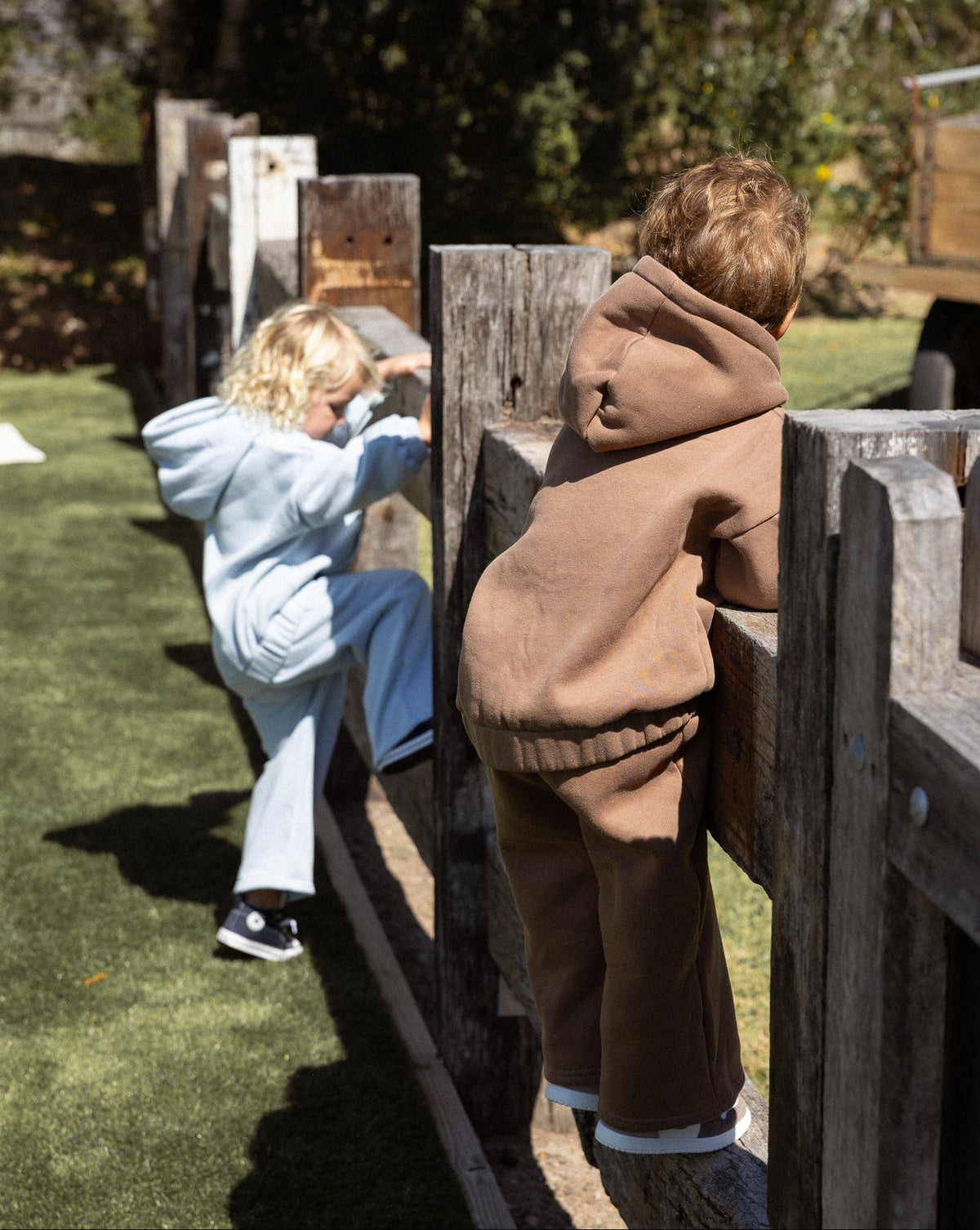 Two children outdoors wearing matching two-piece tracksuits, with one child in the foreground wearing a brown hoodie and pants set and climbing a wooden fence, while another child in a light blue set is visible behind the fence on grass.