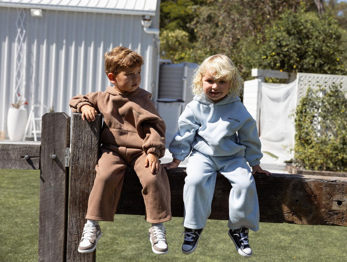 Two children sitting on a wooden fence outdoors, wearing matching oversized tracksuits. The child on the left wears a brown set, and the child on the right wears a light blue set.